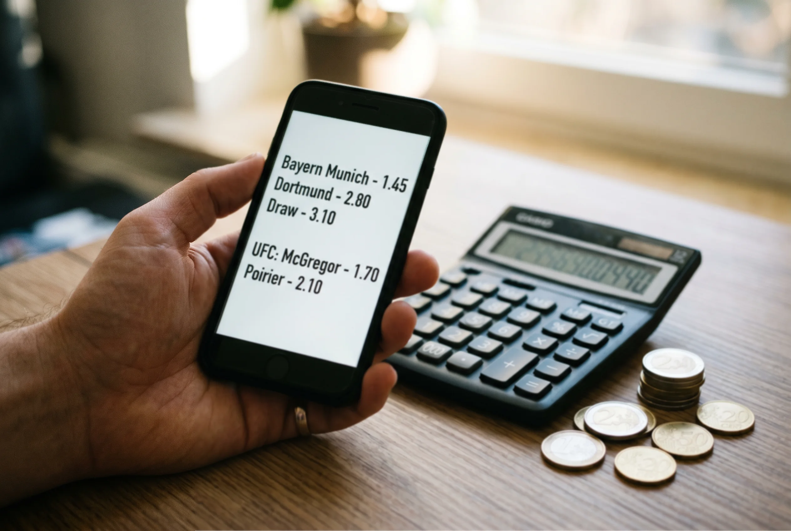 Smartphone mit Wettquoten neben Taschenrechner und Euro-Münzen auf einem Schreibtisch